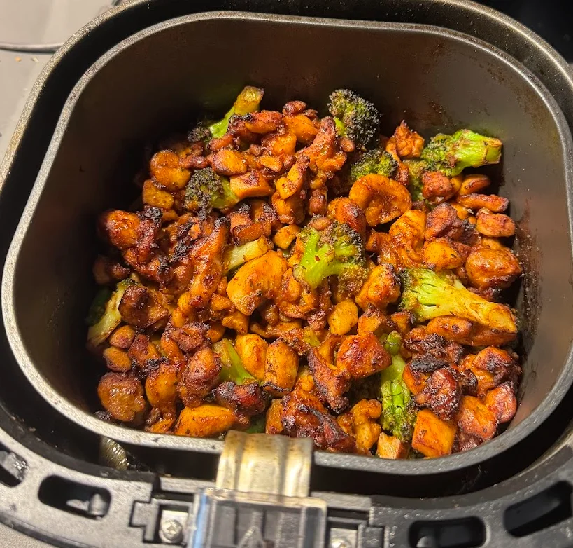 Browned pieces of chicken thigh and broccoli in an open air fryer basket.